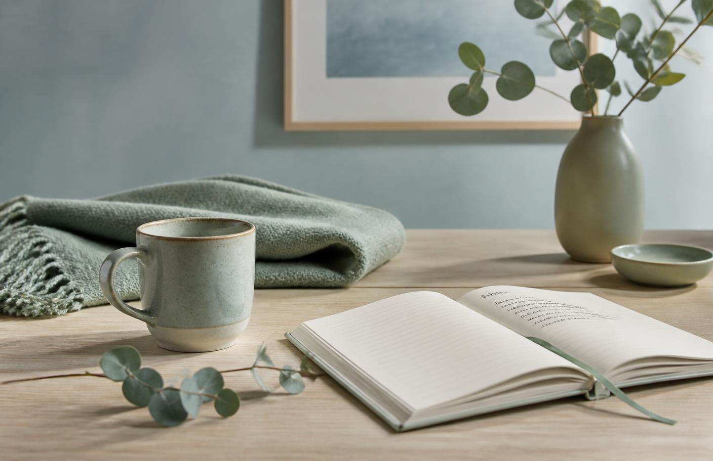 An open notebook and ceramic mug beside a folded throw and green branches